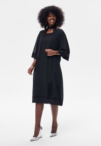 Kobieta o kręconych włosach, ubrana w czarną sukienkę do kolan, dopasowany szalik i srebrne szpilki, stojąca i uśmiechająca się na jednolitym tle.