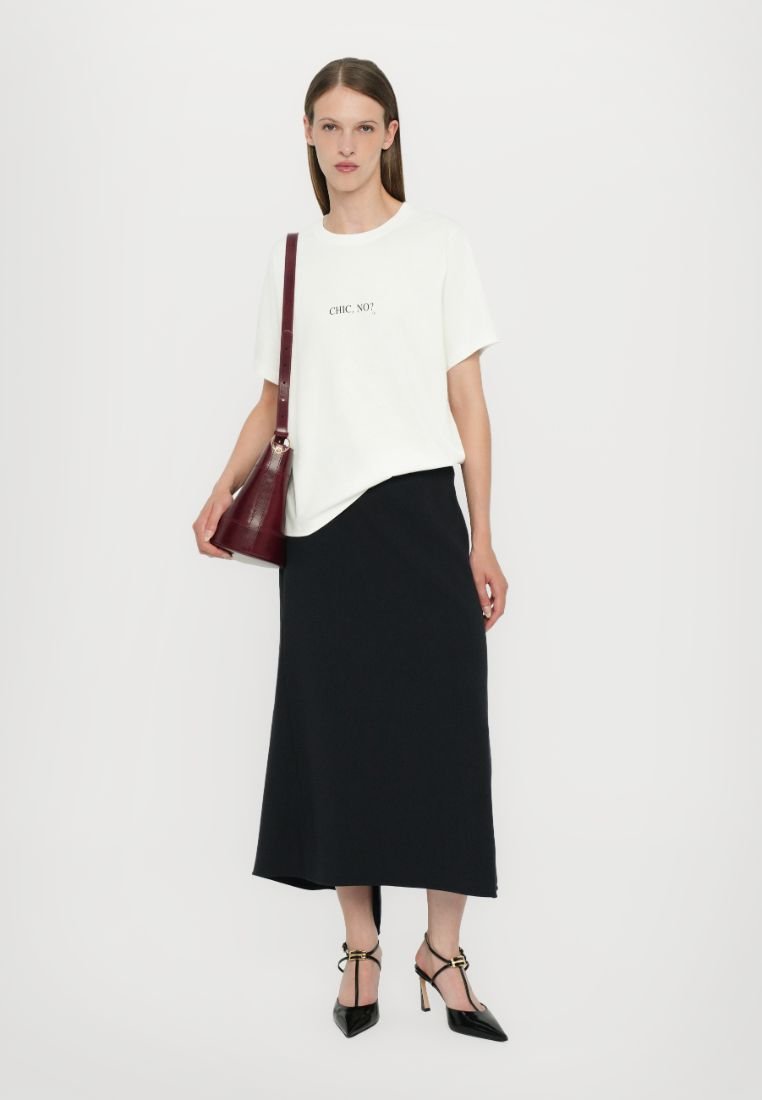 T-shirt blanc avec le texte "CHIC, NON ?" associé à une jupe midi noire. Les accessoires incluent un sac bourgogne et des chaussures à talons noires avec des brides aux chevilles.
