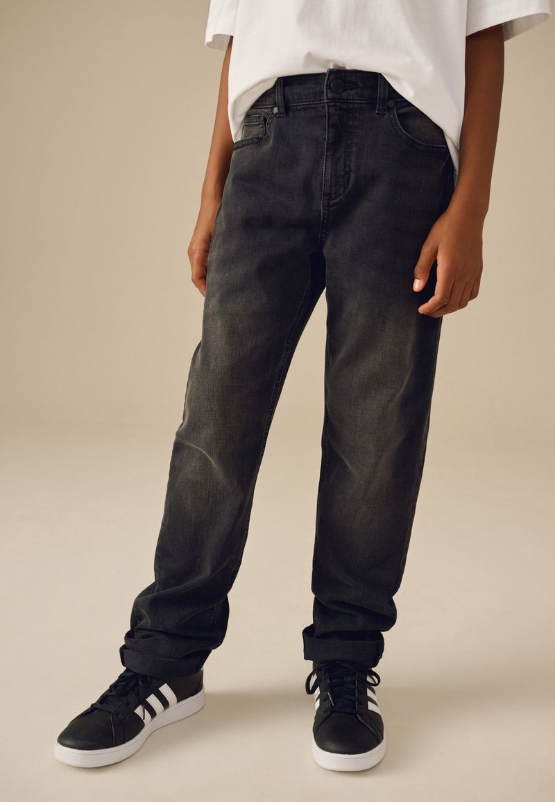 Personne portant un jean noir délavé à jambes droites, un t-shirt blanc et des baskets noires avec des bandes blanches, debout devant un fond beige uni.