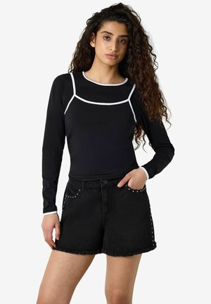 Mujer con cabello largo y rizado que lleva una camiseta negra de manga larga con ribete blanco y pantalones cortos de mezclilla negros con tachuelas metálicas.