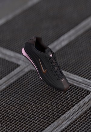 Schwarzer und dunkelbrauner Nike-Sneaker mit pinker Sohle, Schnürsenkel gebunden, diagonal auf einer Metallgitteroberfläche platziert.