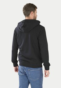 Hoodie en tissu noir avec capuche à cordons, poignets et ourlet côtelés. Porté avec un jean en denim bleu, vu de dos.