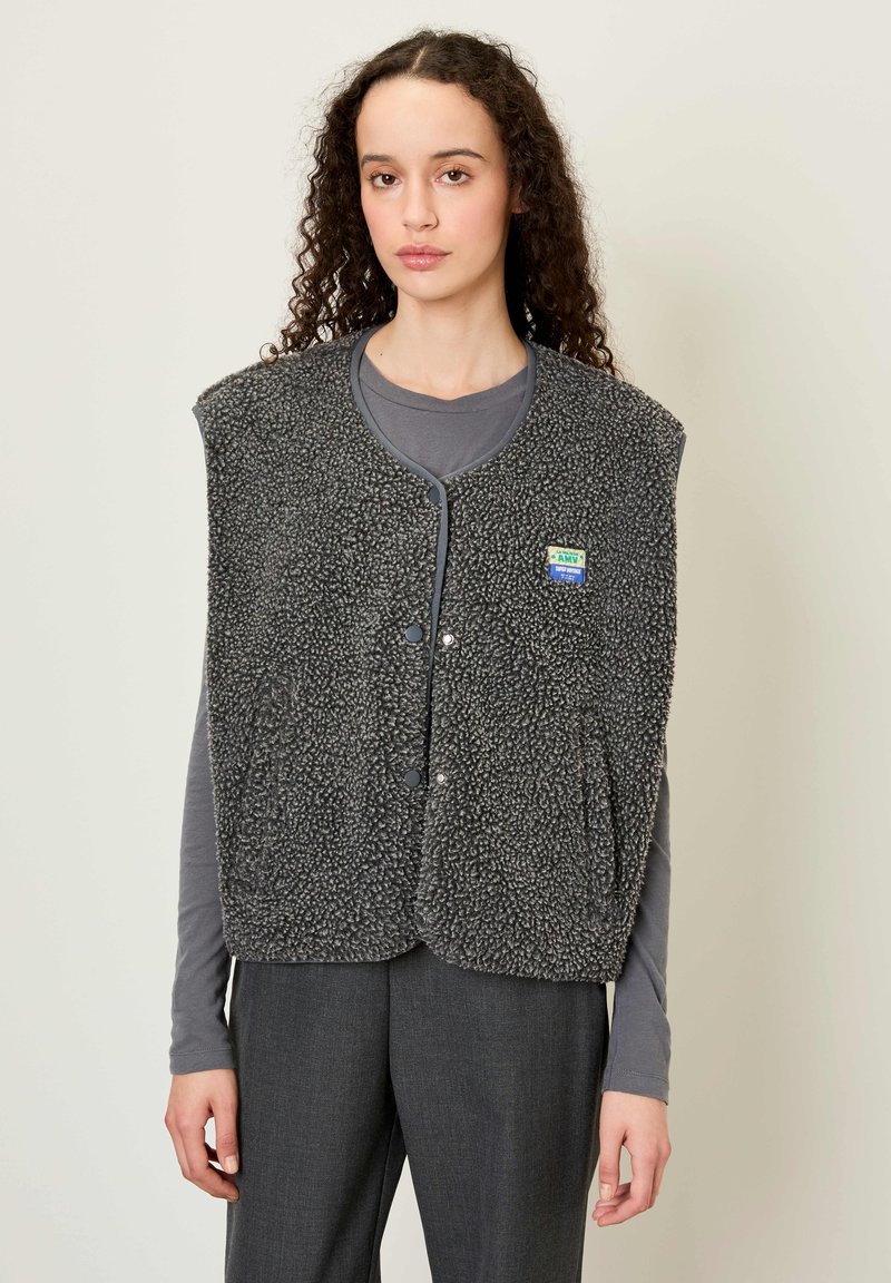 Pelēkā teddy vilnas veste ar noapaļotu kakla izgriezumu, pogu aizdari un sānu kabatām. Valkāta virs pelēka garām piedurknēm krekla, minimālisma dizains.