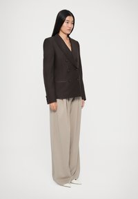 Blazer marrón de doble botonadura con solapas con muesca, combinado con pantalones beige holgados y zapatos blancos puntiagudos. Texturas suaves en todo.