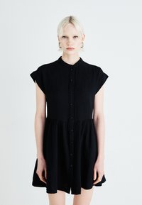 Rochie neagră cu mâneci scurte, guler mandarin și fustă evazată. Confecționată dintr-un material texturat. Fără modele sau accesorii.