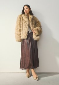 Brūns mākslīgais kažoks ar platām piedurknēm, uzlikts virs tumša plisēta svārka. Skaidri redzami zelta norādītās papēža kurpes ar loku akcentiem.