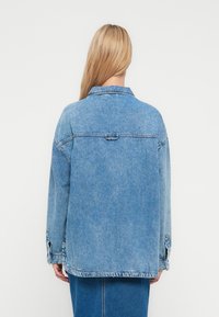 Light blue denim jacket, relaxed fit with a collar, visible stitching, and buttoned cuff details. Features a centered back seam.
