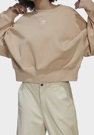 Personne portant un sweat-shirt Adidas beige oversize avec logo et un pantalon taille haute crème clair, les mains touchant les cheveux et les côtés du sweat-shirt.