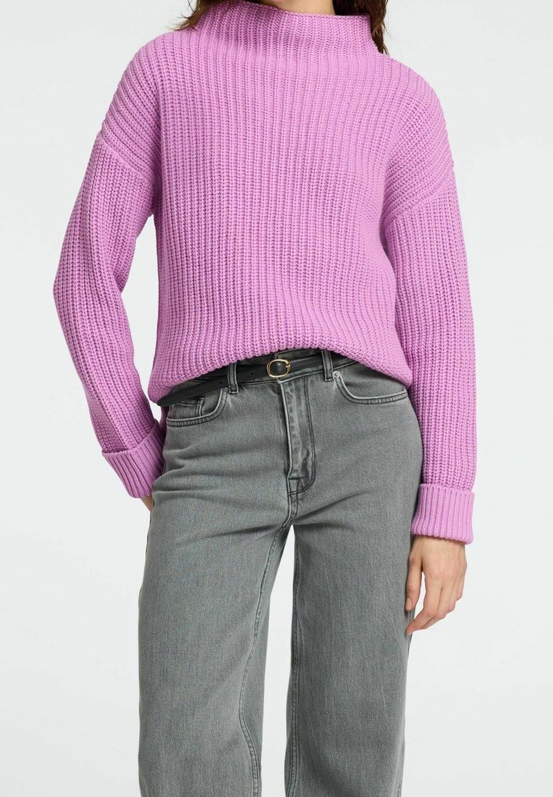 Pull rose tricoté à col montant et texture côtelée, associé à un jean gris taille haute avec une ceinture en accent.