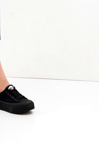 Juodas platformos sportbačio batelis ant žmogaus kojos baltame fone, matomas šoninis vaizdas su raišteliais ir tekstūruota padais.