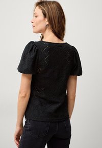 Schwarzes Spitzenoberteil mit kurzen Puffärmeln, das mit einem strukturierten Blumenmuster und einem runden Ausschnitt versehen ist, kombiniert mit dunklen Jeans.