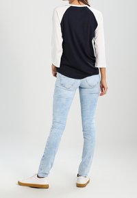 White and navy baseball tee with three-quarter sleeves, paired with light blue skinny jeans and white sneakers. Back view shown, seamless texture.