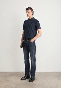 Námornícky polo tričko s krátkymi rukávmi a golierom, spárované s tmavými džínsami. Čierne topánky dotvárajú outfit. Minimálne značenie na tričku.