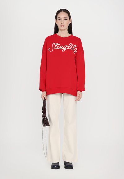 Sudadera roja de gran tamaño con el texto blanco "Steiglitz" en la parte delantera, combinada con pantalones de pierna ancha color crema y zapatos oscuros, sosteniendo un bolso pequeño.