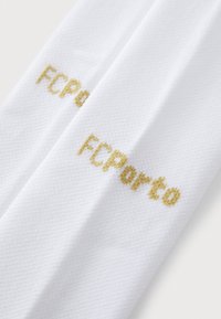 Bela tkanina športnih nogavic v beli barvi z zlatim napisom "FC Porto". Napis je izvezen v kockastem fontu. Tekstura se zdi gladka in zračna.