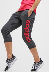 Donkergrijze cropped sweatpants met een verticaal gedrukt rood "adidas" logo op het linkerbeen. De stof lijkt zacht en heeft een comfortabele pasvorm.