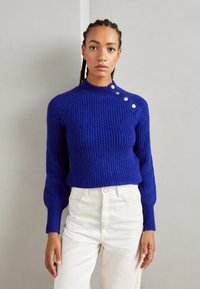 Royal blue ribbed knit sweater with a high neck and button detail on the shoulder, paired with white high-waisted pants.