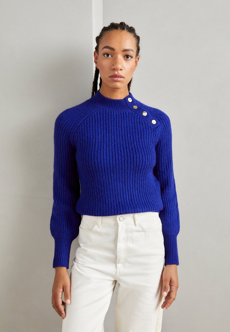 Pull en maille côtelée bleu royal avec un col montant et un détail de bouton à l'épaule, associé à un pantalon blanc taille haute.
