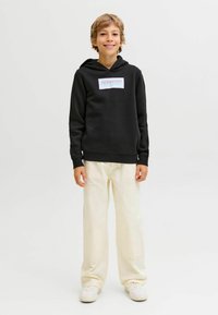 Sudadera negra con un logo frontal en blanco y rojo, combinada con pantalones anchos de color crema y zapatillas blancas. Diseño simple, corte informal.