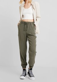 Olivgröna joggerbyxor med elastiska muddar och justerbart snöre i midjan, kombinerat med en vit croppad topp och svarta hög-top sneakers.