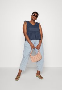 Top sem mangas azul marinho com barra peplum e detalhes de laço ajustáveis nos ombros, combinado com jeans baggy claros e sandálias verdes.
