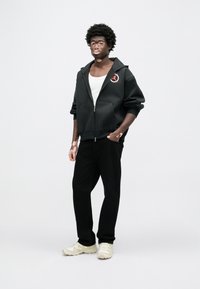 Μαύρο φούτερ με φερμουάρ και λογότυπο, φορεμένο πάνω από λευκό αμάνικο τοπ. Συνδυασμένο με μαύρο παντελόνι και ανοιχτόχρωμα αθλητικά παπούτσια. Casual εφαρμογή και σχέδιο.