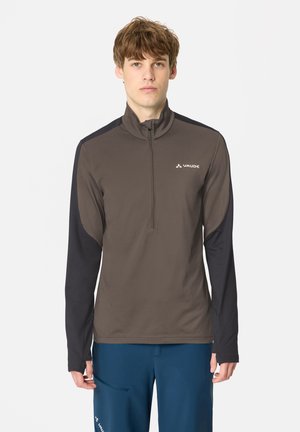 Giovane uomo con capelli castani corti che indossa una maglia marrone e nera a maniche lunghe con zip lunga un quarto e pantaloni blu, in piedi su uno sfondo chiaro.