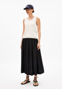 Cremfarbener, gestrickter ärmelloser Pullover mit V-Ausschnitt, schwarer, plissierter Maxi-Rock und schwarze, mit Riemen umwickelte Sandalen mit Perlen. Dunkle Kappe auf dem Kopf.