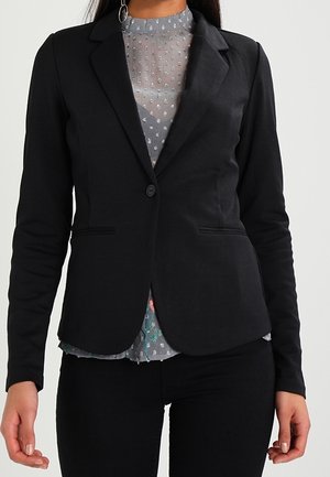 Zwarte op maat gemaakte blazer met een enkele knoopsluiting, voorzien van een revers en twee zakken aan de voorkant. Daaronder bevindt zich een grijze geblokte blouse.