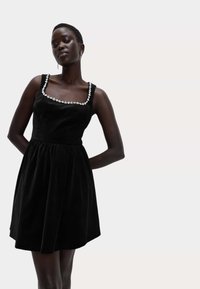 Černé sametové šaty s přiléhavým korzetem, čtvercovým výstřihem a zdobeným květinovým lemováním. Rozšířená sukně sahá nad kolena.