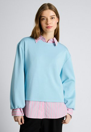 Jonge vrouw met lang lichtbruin haar, gekleed in een lichtblauwe oversized trui over een roze gestreept shirt en zwarte broek, staand tegen een effen muur.