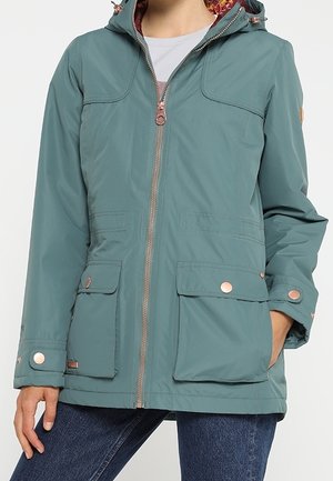 Kvinde iført en teal hættejakke med forlynlås og klaplommer, kombineret med blå jeans, stående med den ene hånd i lommen.