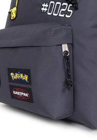 Zaino in tessuto grigio con un patch di Pikachu, ricamato con "#0025" e doppio logo: Pokémon ed Eastpak su etichette testurizzate. Include tiralampo.