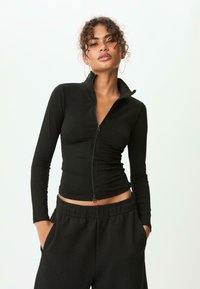 Femme aux cheveux bouclés portant un haut noir à fermeture éclair et un pantalon noir à taille élastique, les mains dans les poches, devant un fond clair et uni.