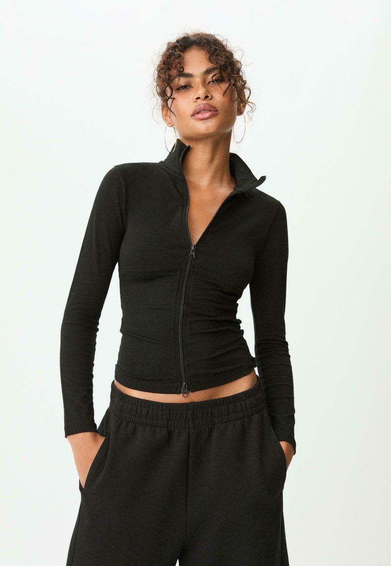 Femme aux cheveux bouclés portant un haut noir à fermeture éclair et un pantalon noir à taille élastique, les mains dans les poches, devant un fond clair et uni.