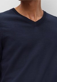 Námořnická modrá pletená svetr s výstřihem do V, s hladkým povrchem a dokončenými švy. Viditelné švy podél výstřihu.