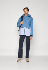 Tânăr îmbrăcat cu o jachetă albastră și gri deschis, pantaloni negri, adidași bej și purtând un rucsac negru pe o umăr.