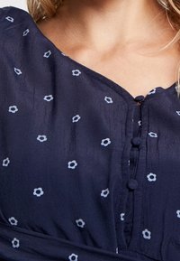 Marinblus med små vita blommönster, V-ringning, knäppning och strukturerad tyg. Subtil drapering vid fållen.