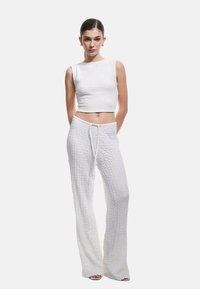 Młoda kobieta z wygolonymi włosami, ubrana w białą, teksturowaną, bezrękawnikową bluzkę crop top oraz pasujące, szerokie spodnie z troczkiem, stojąca na białym tle.