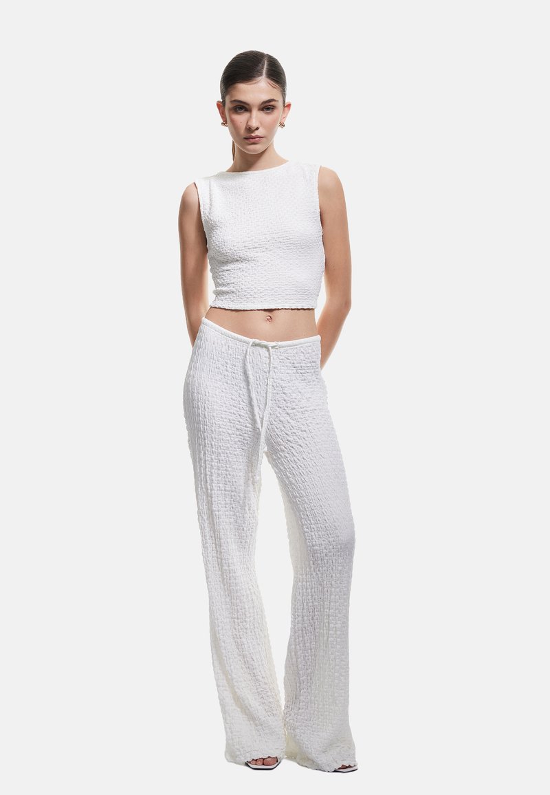 Młoda kobieta z wygolonymi włosami, ubrana w białą, teksturowaną, bezrękawnikową bluzkę crop top oraz pasujące, szerokie spodnie z troczkiem, stojąca na białym tle.