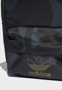 Black fabric backpack featuring a subtle camo pattern, zip closure, front pocket with a gold Adidas logo, and textured surface.