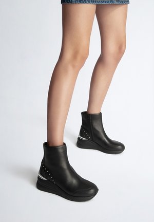 Jambes portant des bottines en cuir noir avec fermetures éclair sur les côtés et détails en clous argentés, associées à une jupe courte en jean sur un fond uni.