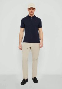Homem de pé, vestindo uma polo azul-marinho, calças bege, sapatos pretos e um chapéu bege, contra um fundo claro e simples.