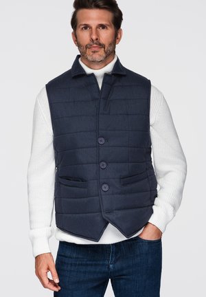 Uomo che indossa un gilet trapuntato blu navy sopra un maglione bianco a maglia e jeans blu scuro, in piedi con una mano in tasca contro uno sfondo semplice.