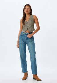 Chaleco corto a cuadros en blanco y negro con cierre de botón, combinado con jeans azules de cintura alta y botas de tobillo en color marrón. Textura de tela suave.