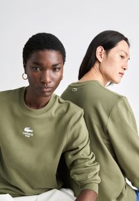 Olivovo zelená mikina vyrobená z jemného materiálu, s dizajnom okrúhleho výstrihu, rebrovanými manžetami a vyšitým logom Lacoste na hrudi a chrbte.