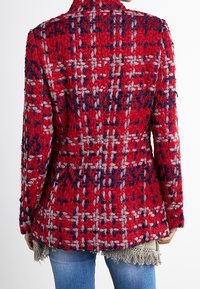 Giacca di lana rossa con una texture a pelo e un motivo a scacchi multicolore, indossata sopra una maglietta beige con frange e jeans blu, vista da dietro.