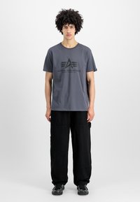 Graues Baumwoll-T-Shirt mit schwarzem Logo und Text, kombiniert mit lockeren schwarzen Cargo-Hosen, in einer einfachen Studio-Umgebung.