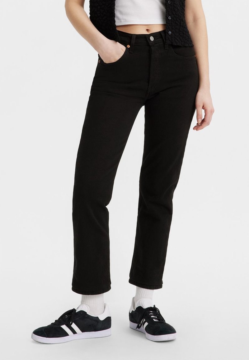 Jeans de mezclilla negra con corte recto, cinco bolsillos, cierre frontal con botón y costuras en contraste. Llevados con zapatillas negras y calcetines blancos.