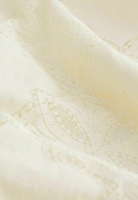 Tissu doux couleur crème avec de délicats motifs en dentelle et des plis subtils créant une texture et des ombres légères.